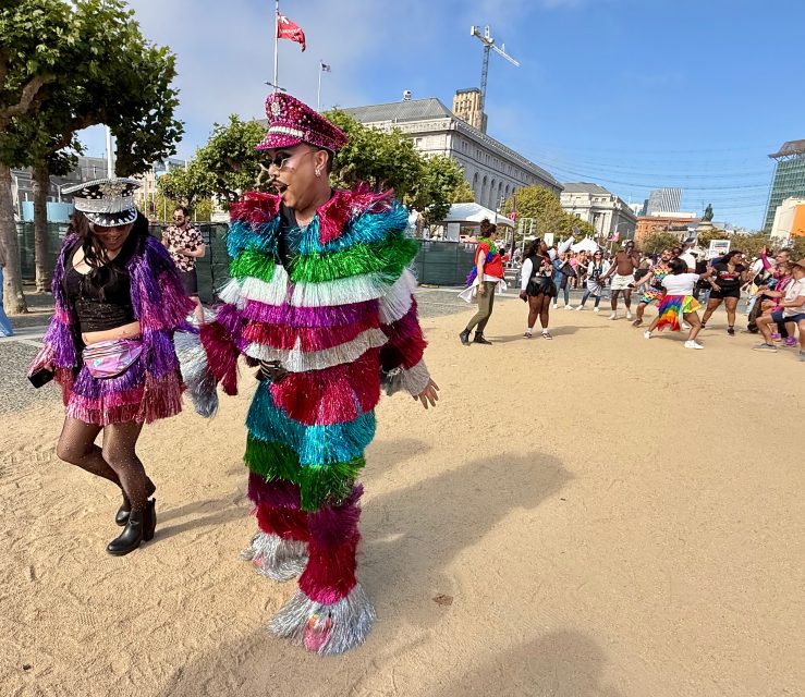 Two people in colorful, fringed costumes walk outdoors during a Pride parade, with a group of people and a large building in the background.