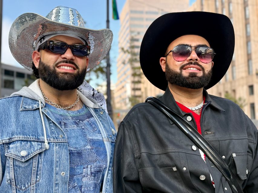 Two men wearing cowboy hats, sunglasses, beards, and Pride vibes pose outdoors in front of a city building; one sports denim overalls and a shiny hat, the other wears a black hat and jacket.