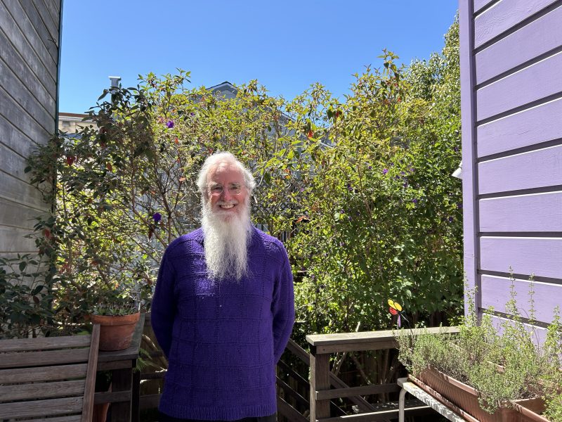 An older man with a long white beard stands outside on a sunny day, wearing a purple sweater, with green bushes and a purple house wall in the background.