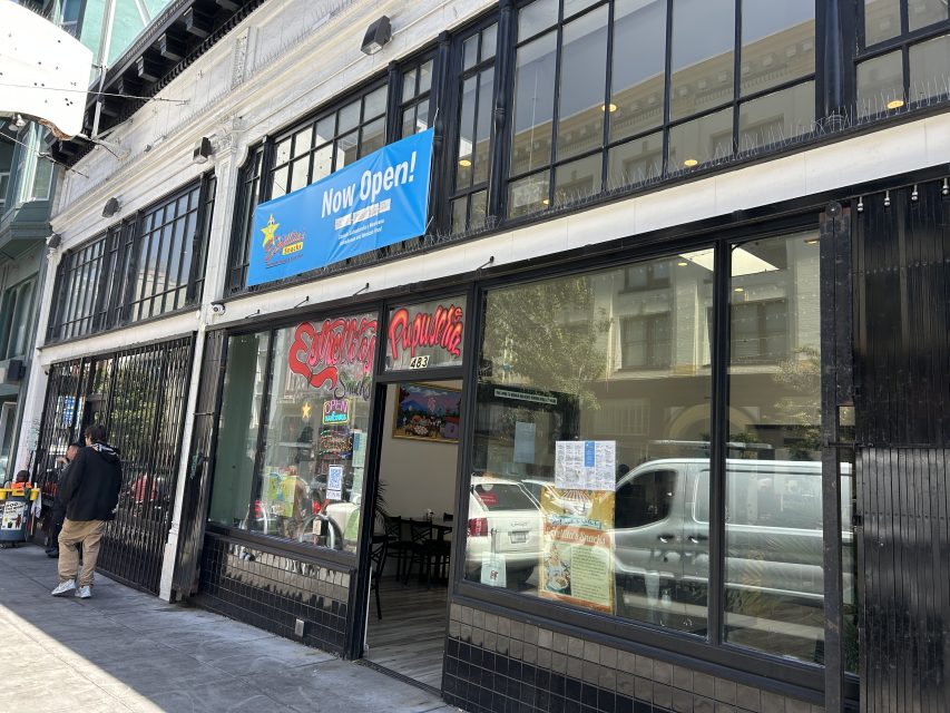 Street view of a storefront with large windows, a blue "Now Open!" banner above the entrance, and a person standing on the sidewalk nearby.