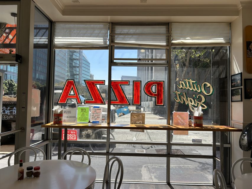 View from inside a pizza restaurant looking out onto the street through large front windows with "PIZZA" painted in red and chairs and tables visible in the foreground.