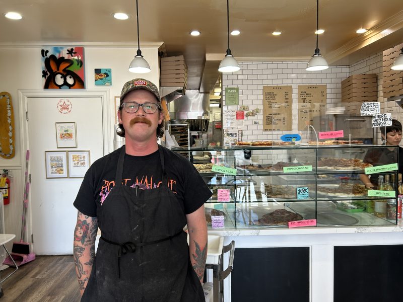 A man wearing a black apron and cap stands in front of a pizza counter inside a brightly lit pizzeria, with pizza menus and display cases visible in the background.