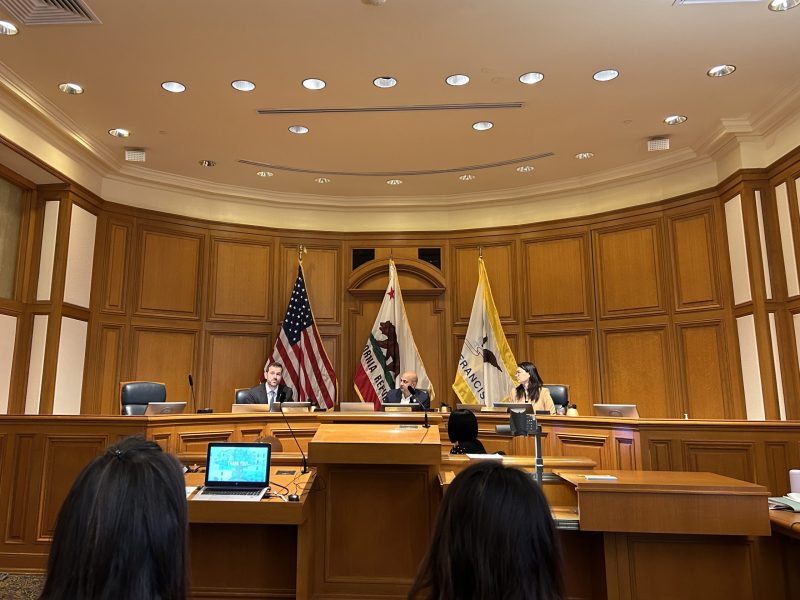A formal courtroom with wooden panels, three people seated at desks, and American, California, and city flags behind them; audience visible in the foreground.