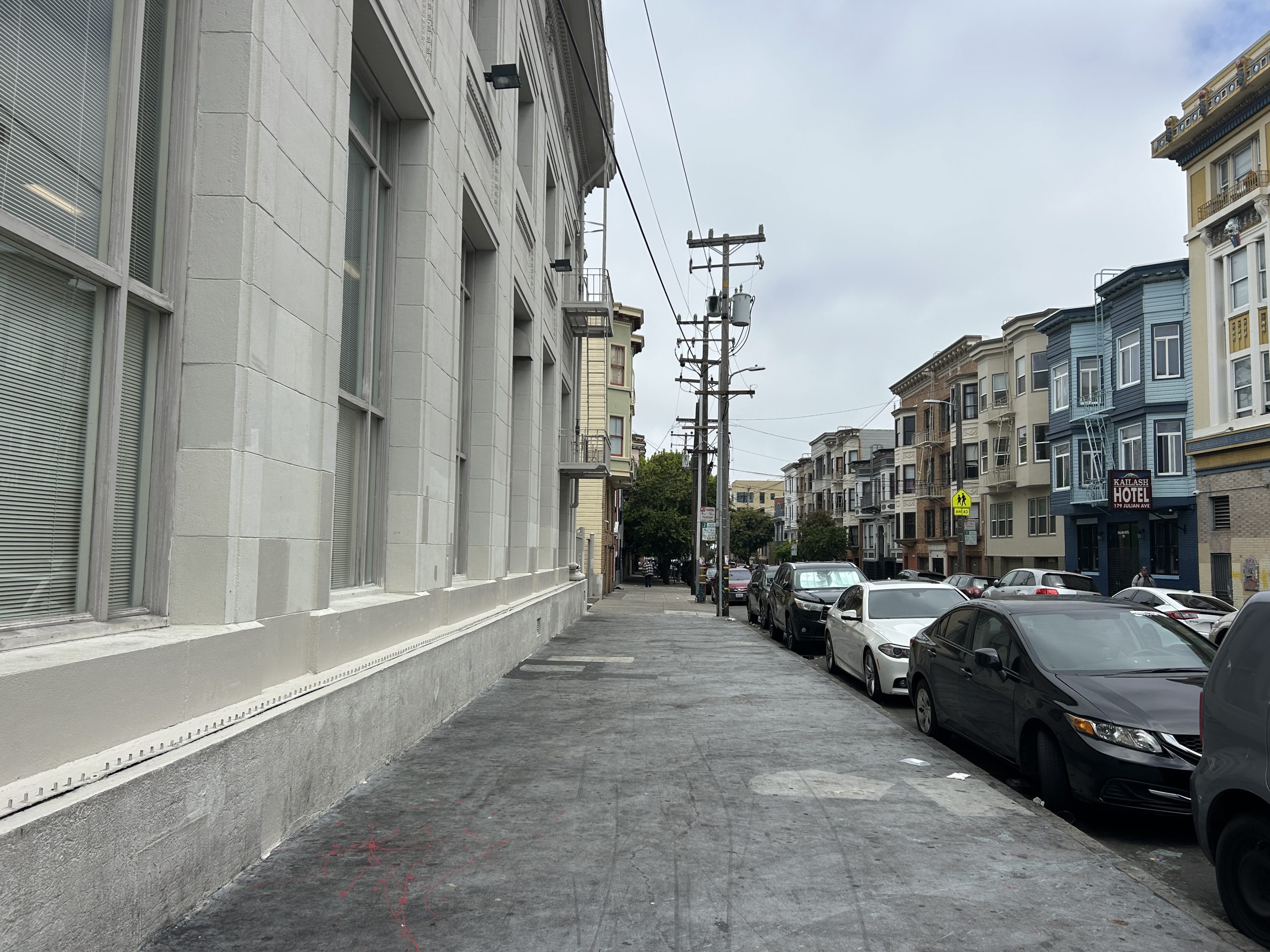 A city sidewalk lined with parked cars, utility poles, and multi-story buildings under a cloudy sky.