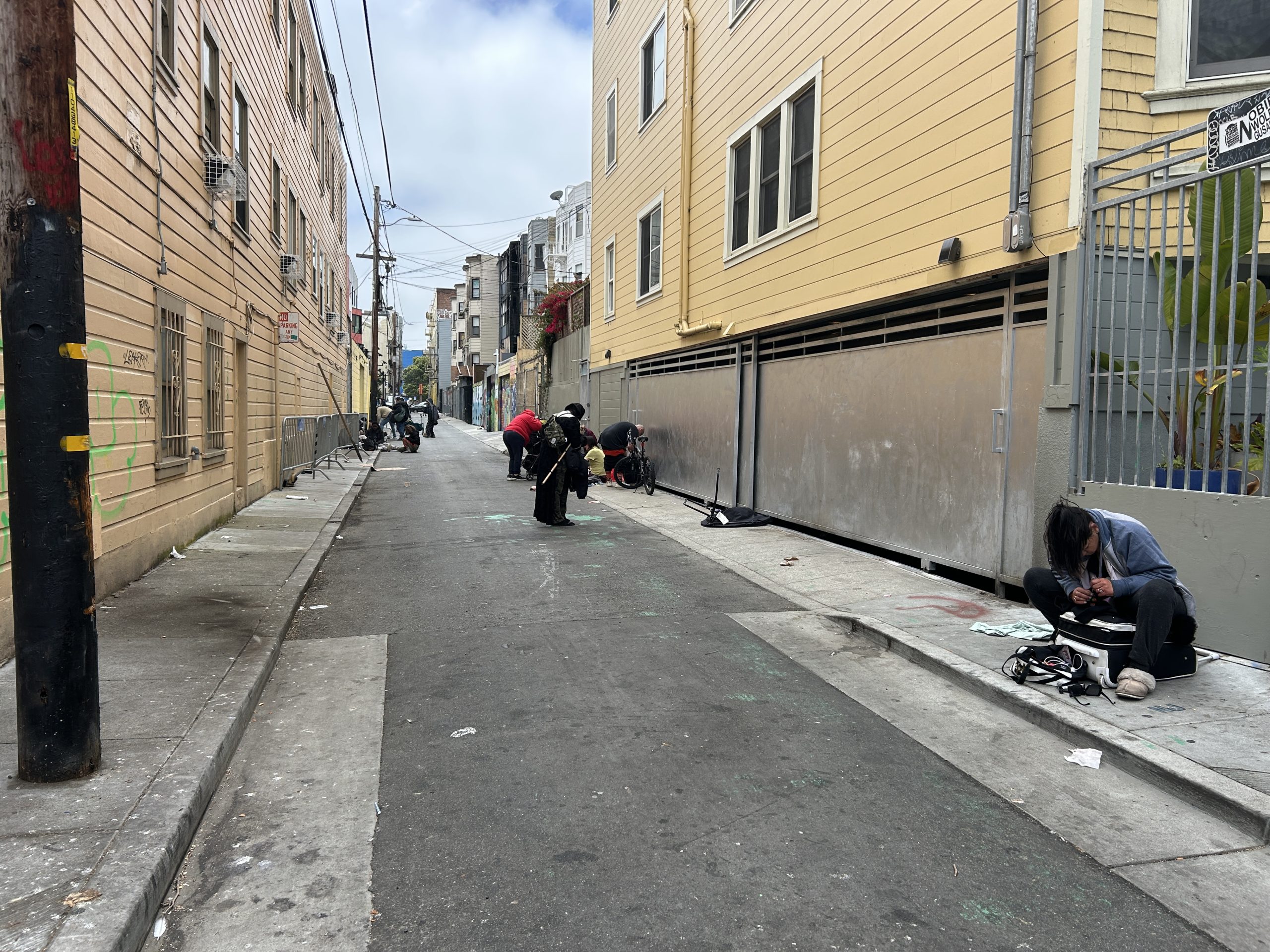 Narrow urban alley with several people sitting or squatting on the sidewalk amid scattered belongings and debris, flanked by multi-story buildings on both sides.
