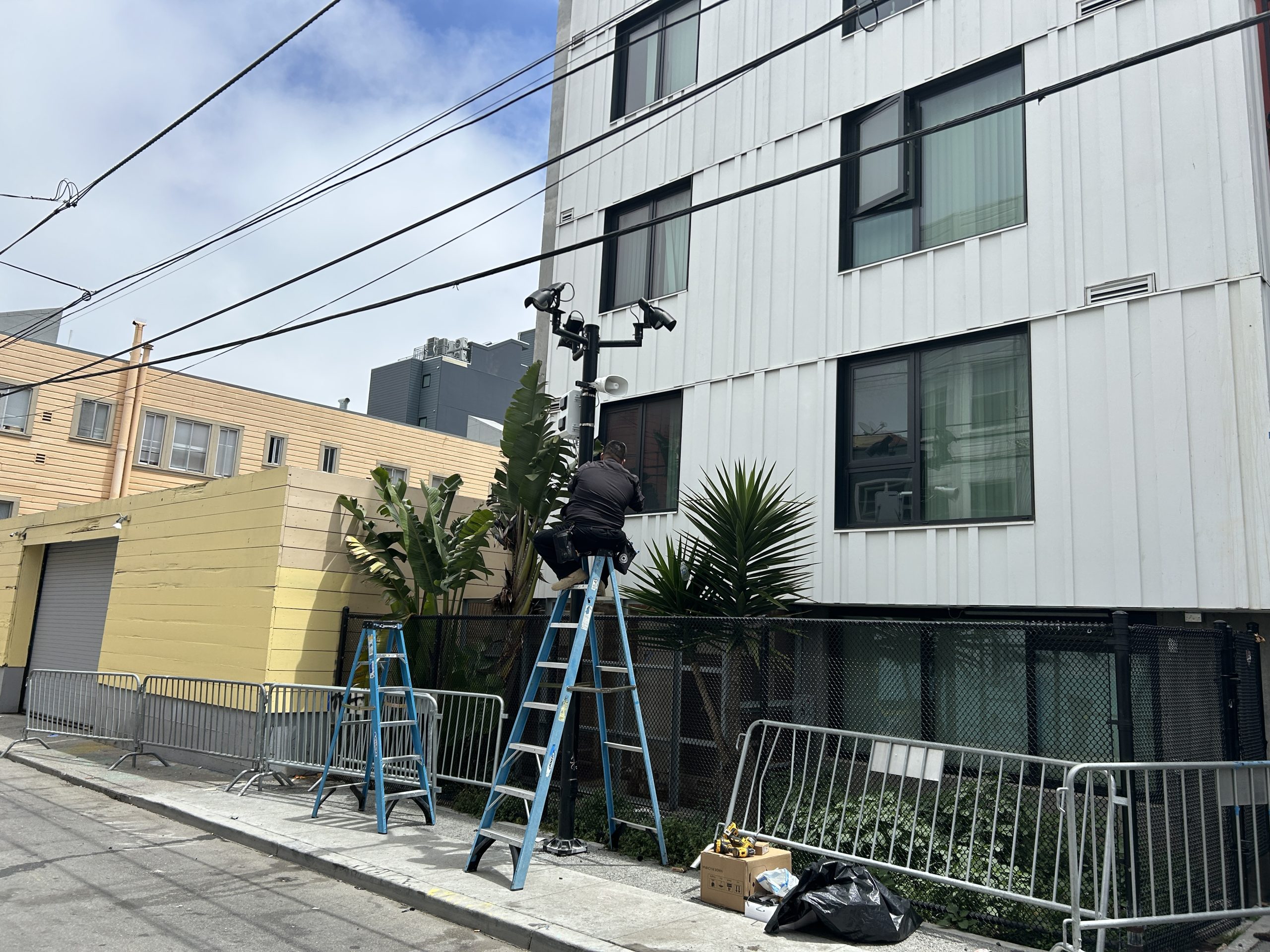 A worker on a ladder installs or repairs security cameras on a utility pole beside a white building, with another ladder and equipment nearby.