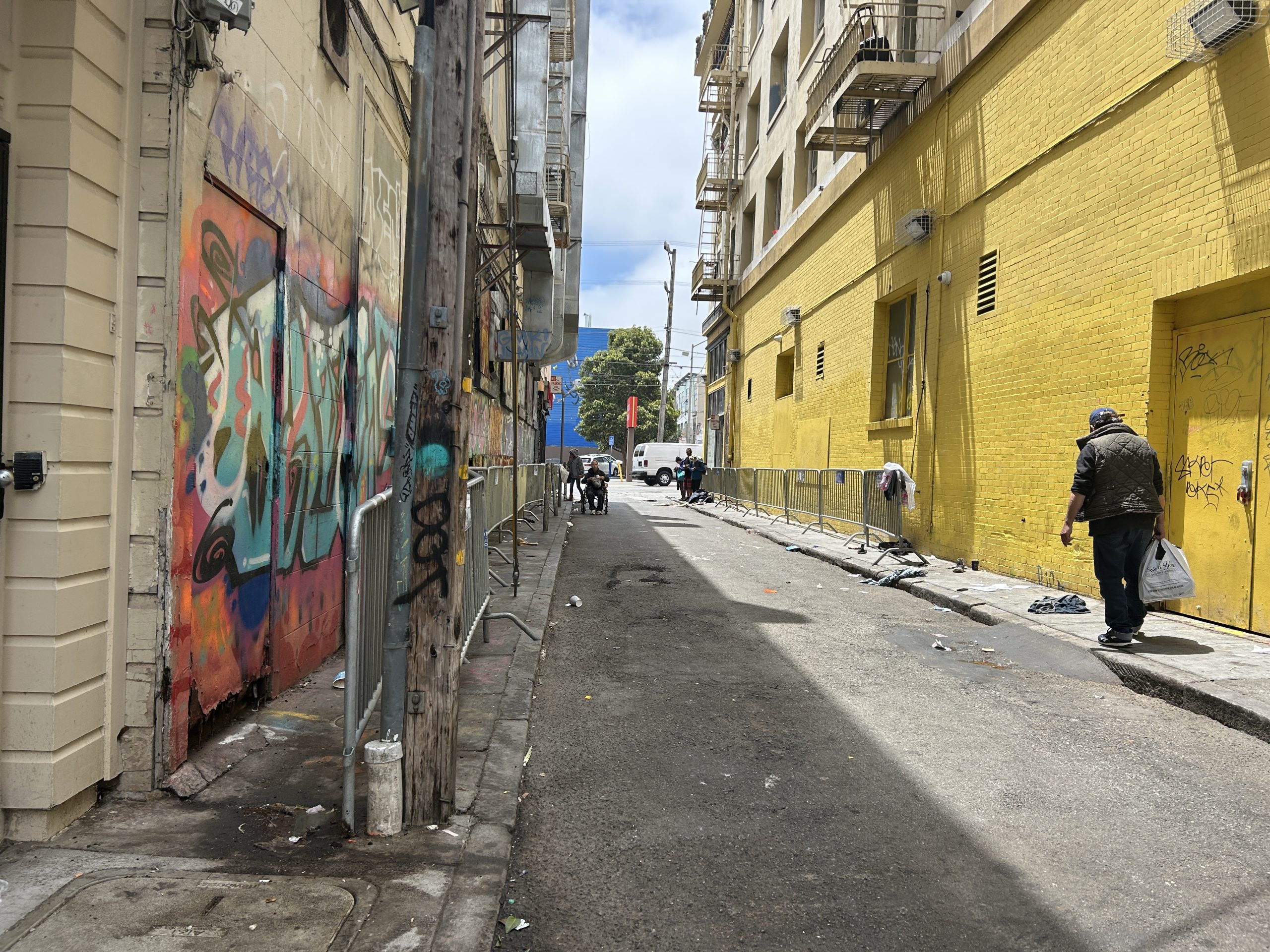 A narrow urban alley with graffiti on one wall, yellow paint on the other, scattered debris, metal barriers, and a person carrying bags walking away.