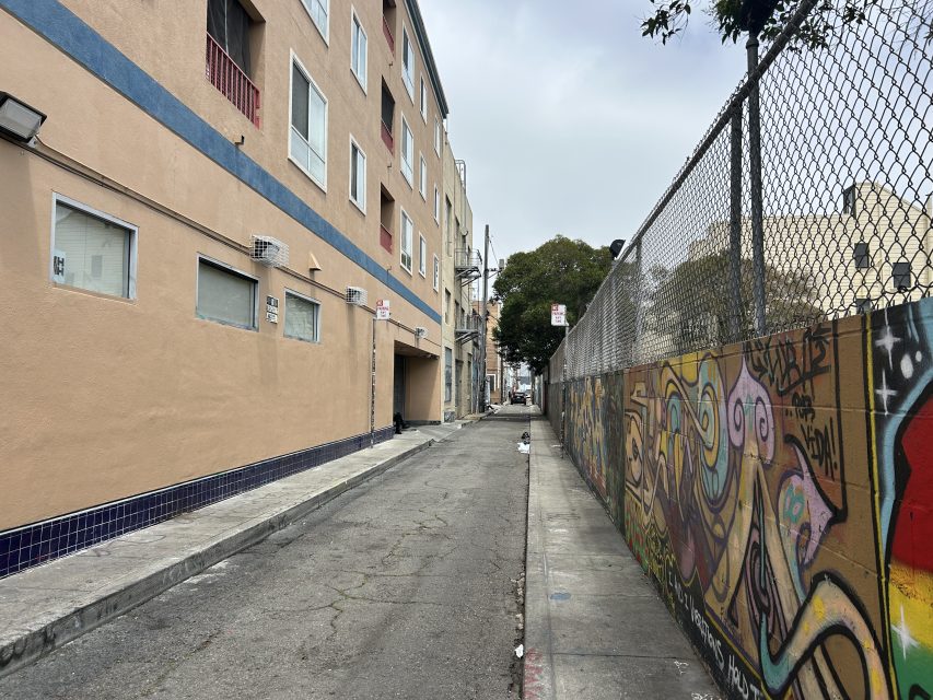 A narrow urban alley with graffiti on a fence to the right, beige apartment building on the left, and a cloudy sky overhead.