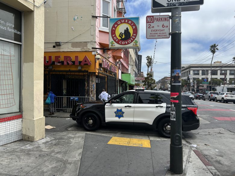 A San Francisco police SUV is parked on the sidewalk outside Taqueria El Coyote, partially blocking a wheelchair ramp at a street intersection.