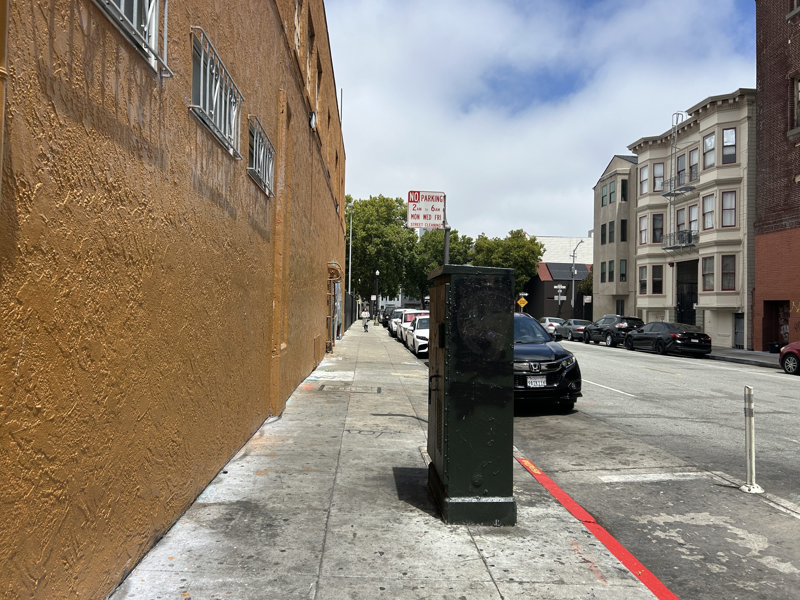 A city sidewalk with parked cars, a utility box, and buildings on both sides under a partly cloudy sky.