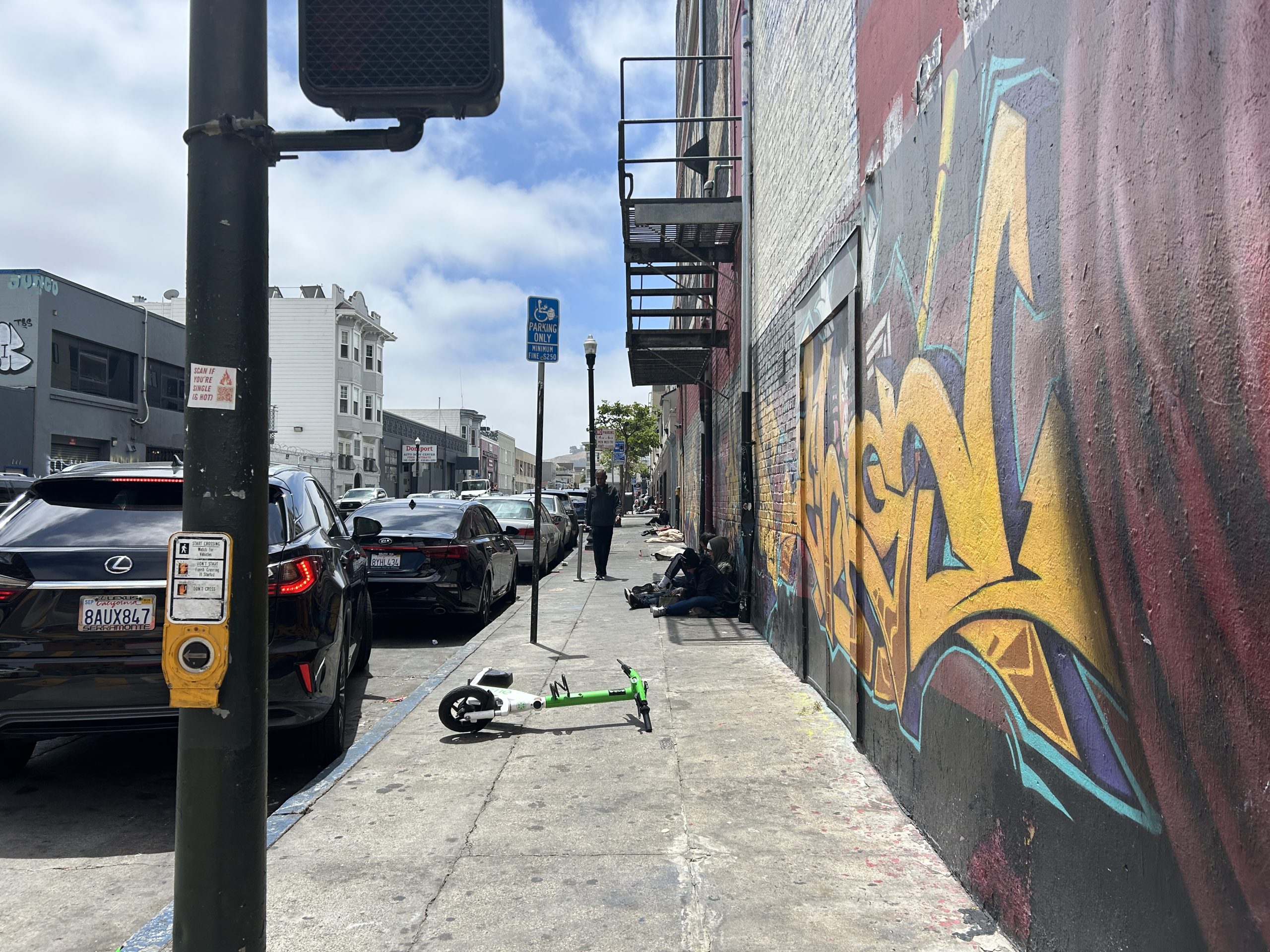 A sidewalk with graffiti on the wall, parked cars, a fallen electric scooter, and people sitting or standing along the building under a partly cloudy sky.