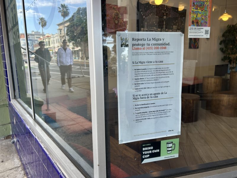 A flyer in Spanish with instructions for reporting immigration enforcement is posted on a business café window; two people are reflected in the glass on a city street.