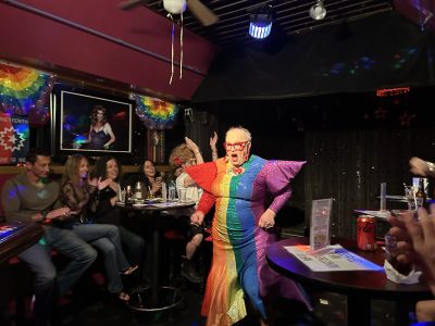 A drag performer in a rainbow gown and bold makeup walks through a lively bar as patrons clap and cheer.