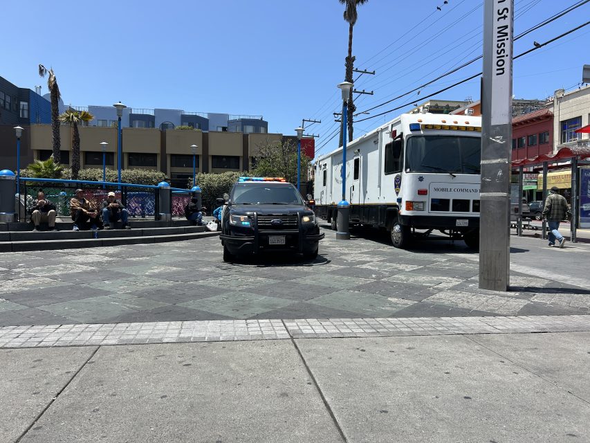 A police car and a large mobile command vehicle are parked near a public plaza, with people sitting and walking nearby on a sunny day.
