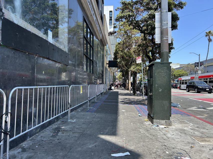 A city sidewalk lined with metal barricades, scattered litter, trees, a traffic signal, and a red and white bus in the street.