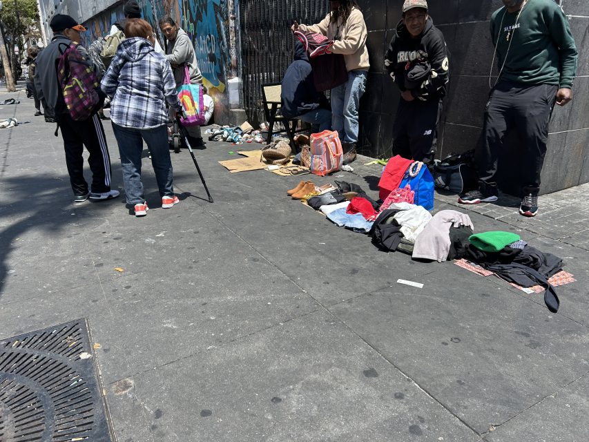 Several people stand and gather on a city sidewalk where clothes and shoes are laid out on the ground, next to bags and backpacks.