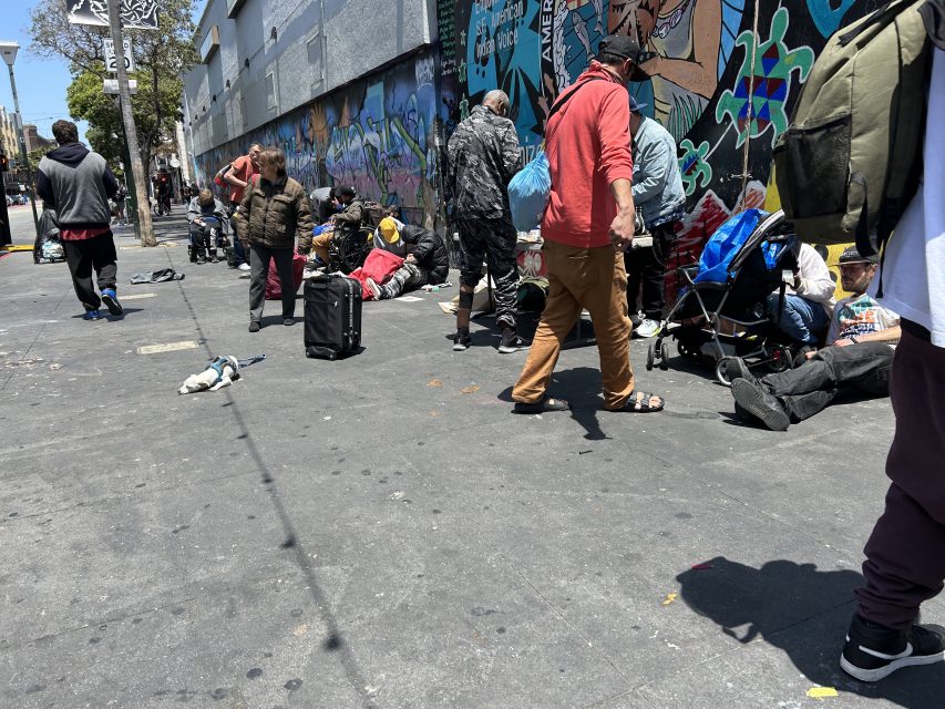 People with belongings gather and sit on a city sidewalk in front of a building with colorful graffiti. Some are standing, some seated, with bags and carts around them.