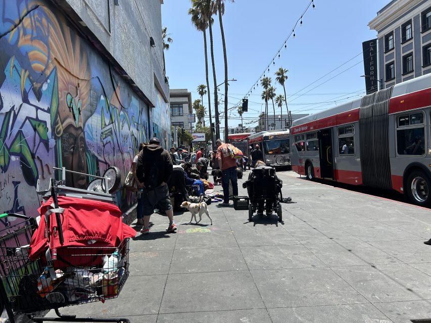 People with belongings gather on a city sidewalk next to a graffiti-covered wall, near a bus stop with buses and palm trees in the background on a sunny day.