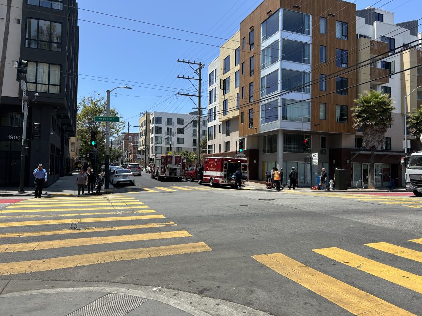 A city intersection with yellow crosswalk lines, apartment buildings, pedestrians, and two fire trucks parked on the street.