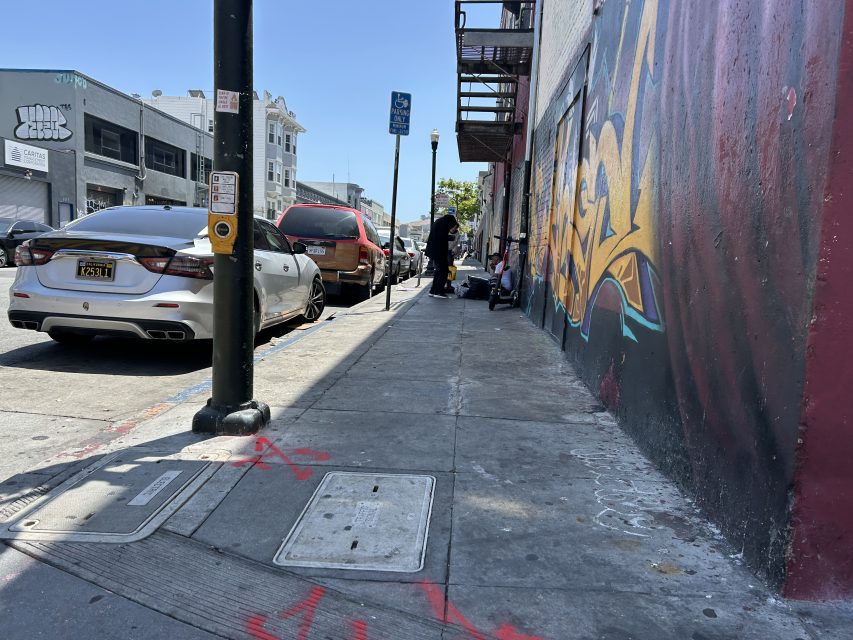 City sidewalk with parked cars on the left, graffiti on the right wall, and a few people sitting and standing near a building under a clear sky.