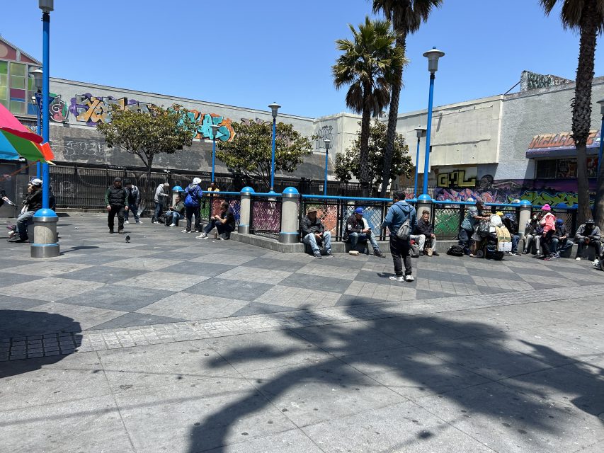 People are sitting and standing in an urban plaza with graffiti-covered walls, trees, and blue lamp posts under a clear sky.