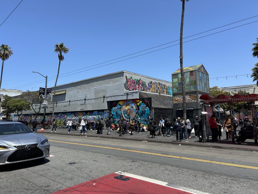 A street scene with people lined up along a sidewalk in front of a building covered in graffiti art, with palm trees and a car passing by in the foreground.