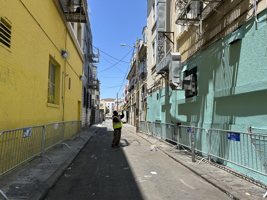 A worker in a safety vest stands in a narrow alley, surrounded by barricades and tall buildings, holding a long pole under a sunny sky.