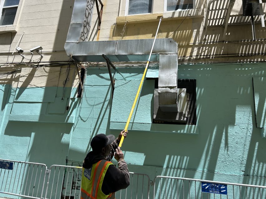 A worker in a safety vest uses an extension pole to paint or clean a high window on a building exterior, with metal ducts and safety barriers visible.