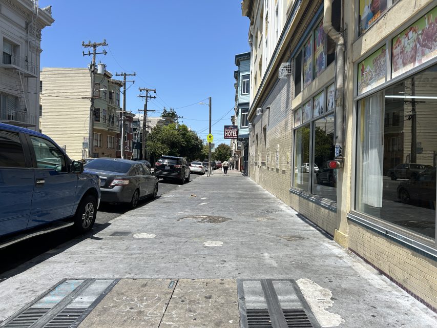 A city sidewalk with parked cars on the left and buildings with shops on the right, under a clear blue sky. A few people walk in the distance.