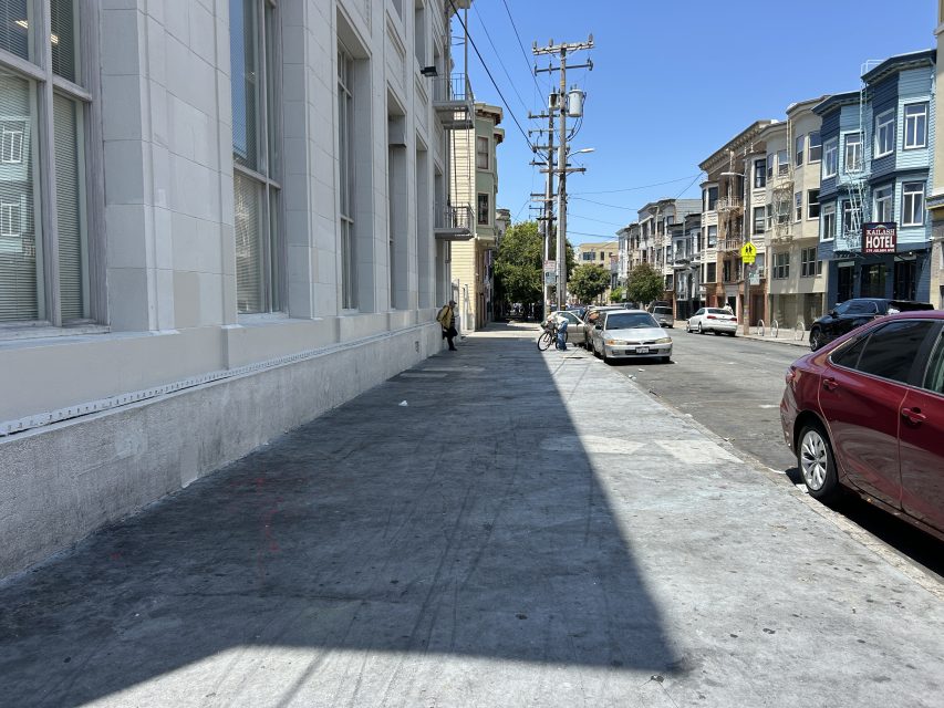 A city sidewalk runs alongside a white building, with parked cars and apartments across the street under a clear blue sky.
