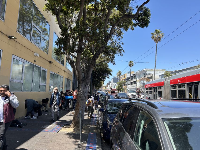 People are gathered on a city sidewalk next to a yellow building and parked cars; a red bus and palm trees are visible in the background under a clear sky.