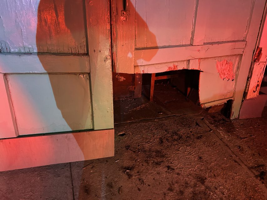 A damaged wooden door with a hole at the bottom and peeling paint, illuminated by red lighting, with a person’s shadow visible on the door.