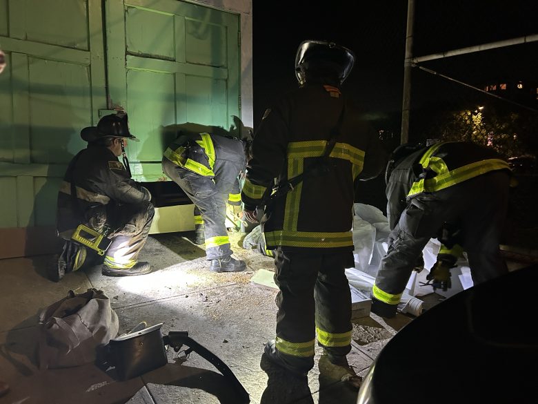 Four firefighters wearing full gear crouch and stand near a building at night, illuminated by flashlights as they examine objects on the ground.