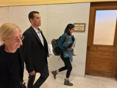 Three people walk down a hallway past a wooden door labeled "Geologist Office." One woman with folders and a backpack appears deep in planning as she walks, while another woman and a man walk side by side.