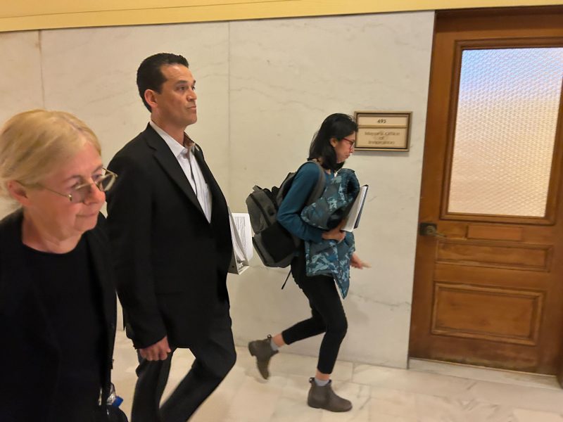 Three people walk down a hallway past a wooden door labeled "Geologist Office." One woman with folders and a backpack appears deep in planning as she walks, while another woman and a man walk side by side.