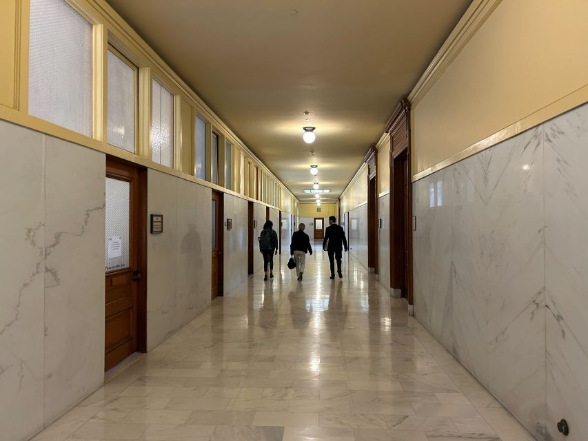A group of four people walk down a long, marble-floored hallway with wooden doors and overhead lights, deep in conversation and Planning their next steps.