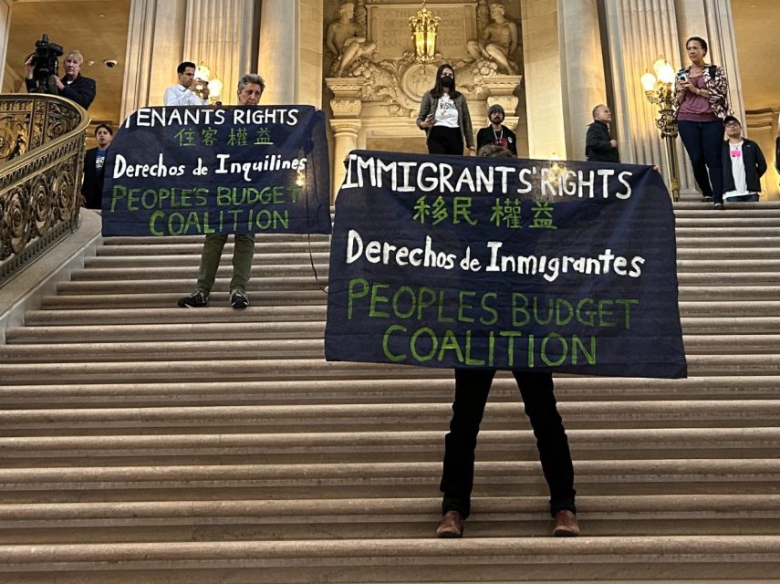 People stand on marble stairs inside a grand building, holding banners that read "Immigrants Rights" and "Tenants Rights" during a protest. Other people observe or walk nearby.