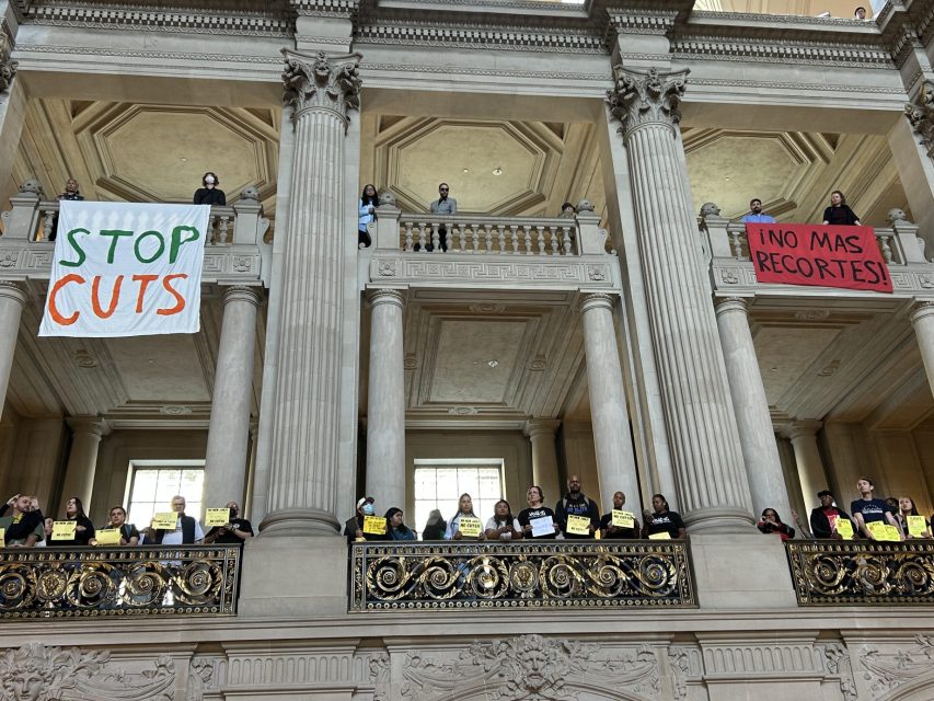 People stand on an ornate balcony inside a government building, holding signs. Two large banners read "STOP CUTS" and "¡NO MÁS RECORTES!".