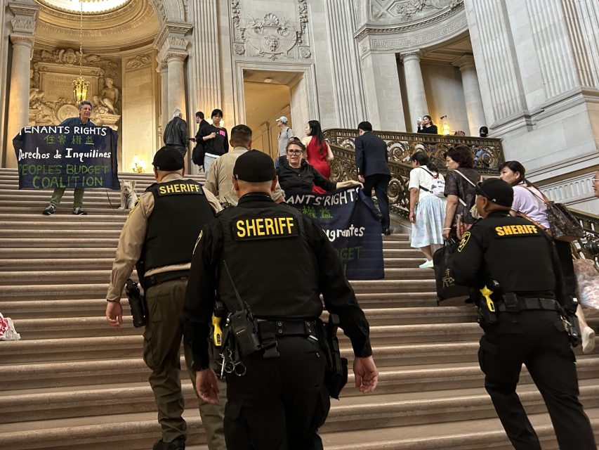 Three sheriffs walk up a grand staircase in a large ornate building toward a group of people holding a banner that reads "IMMIGRANTS RIGHTS.