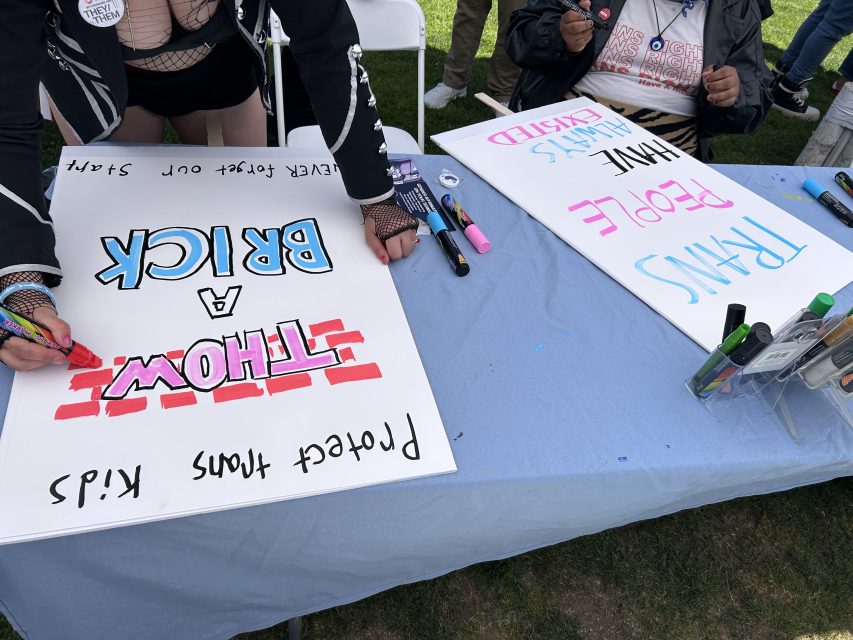 Two people at a table create protest signs with markers. One sign reads "THROW A BRICK," and another reads "TRANS PEOPLE HAVE ALWAYS EXISTED." Art supplies are scattered on the table.
