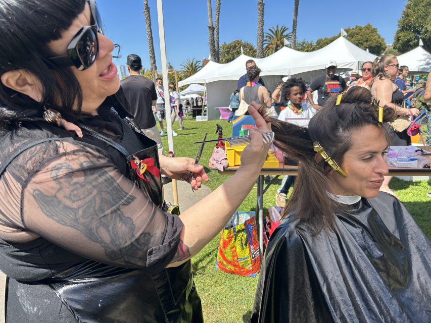 A hairstylist cuts a woman's hair outdoors at an event, with people, tents, and palm trees visible in the background.