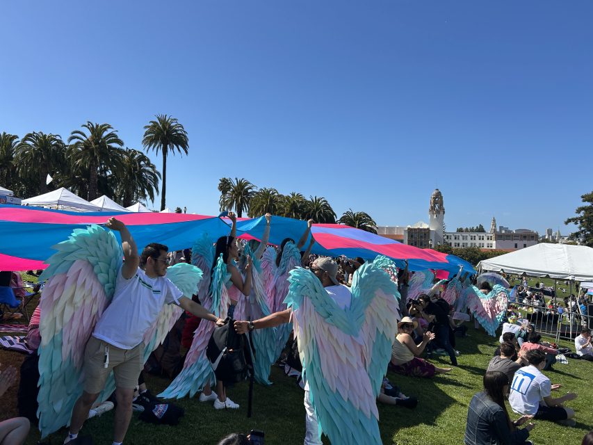 People with pastel angel wings hold up a large transgender pride flag during an outdoor event on a sunny day, with palm trees and buildings in the background.