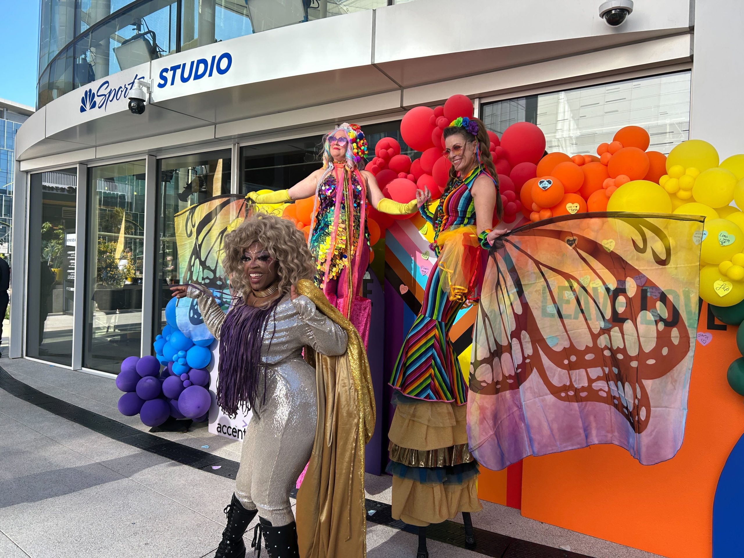 Three people in colorful costumes, including two on stilts with butterfly wings, pose in front of a rainbow balloon display outside an NBC Sports Studio building.