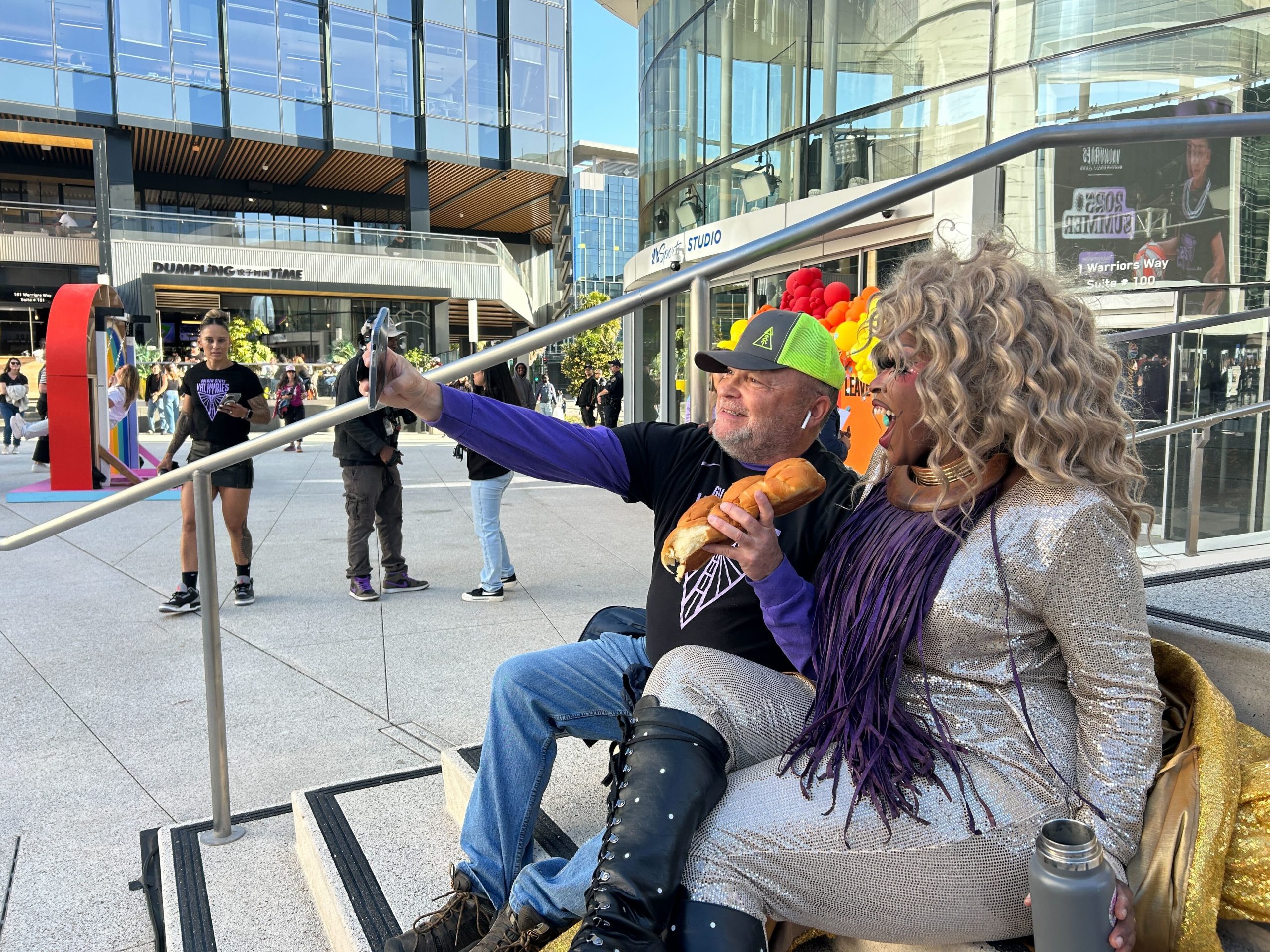A man and a person in a silver outfit sit on steps outdoors, taking a selfie; the latter holds a sandwich. People walk in the background near modern buildings.