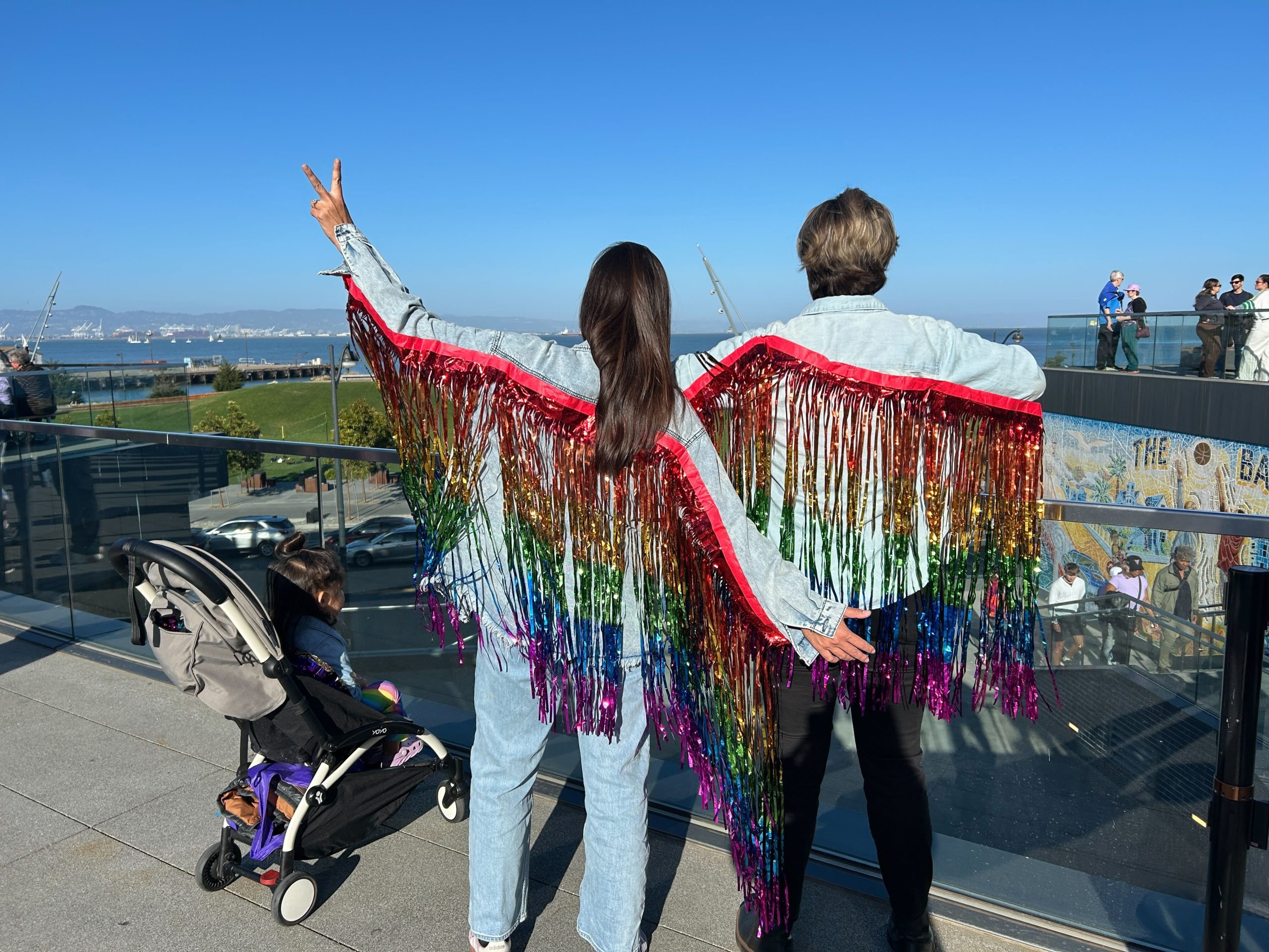 Two people wearing rainbow fringe wings stand side by side, one making a peace sign, while a child in a stroller sits nearby on a sunny rooftop with city and water views.