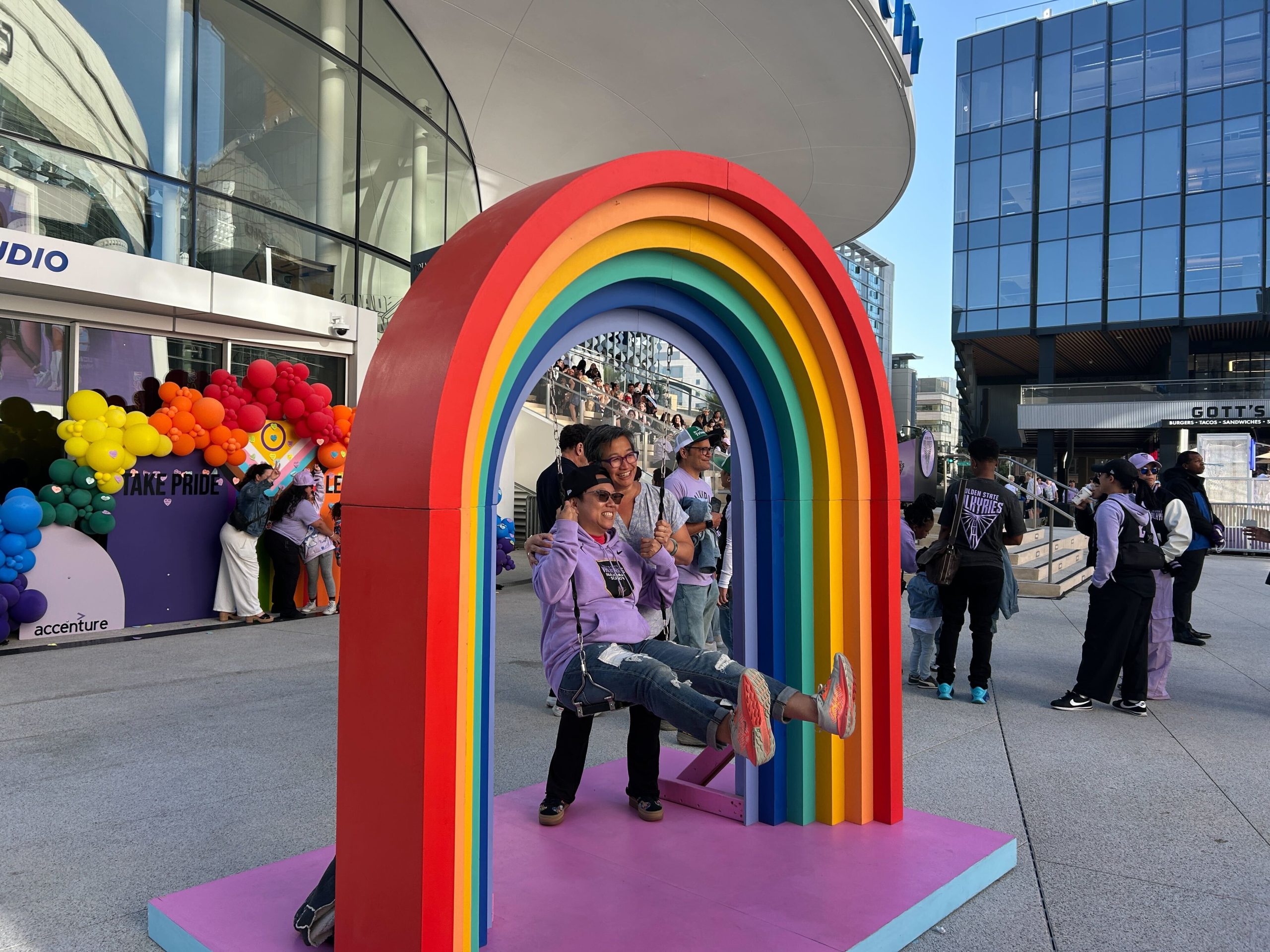 People pose and smile on a swing installed under a large rainbow arch at an outdoor event, with a crowd and balloon decorations in the background.
