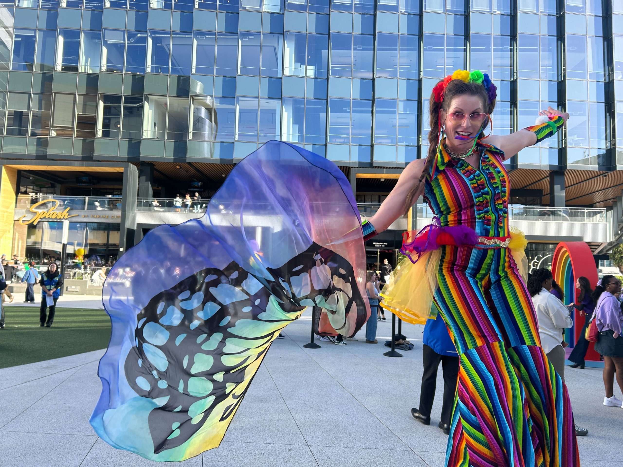 A person in a rainbow-striped outfit and butterfly wing shawl stands on stilts in front of a modern glass building during an outdoor event.