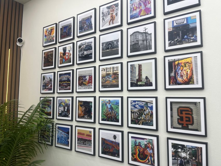 A grid of framed photos and art pieces featuring urban scenes and murals is displayed on a white wall, with a green plant visible in the lower left corner.