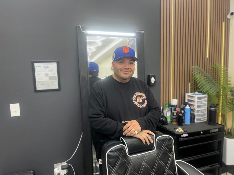 A man wearing a black sweatshirt and blue cap stands behind a barber chair in a modern barbershop, with grooming supplies and a plant visible in the background.