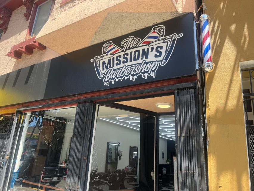 The exterior of The Mission's Barbershop, showing its black sign with barber poles and an open glass-front entrance with a classic barber pole mounted on the wall.